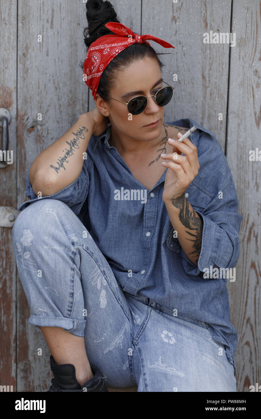 Apropiado Terraplén refugiados Elegante y hermoso de América hipster mujer en jeans de moda en ropa con un  pañuelo rojo y gafas de sol de fumar un cigarrillo y sentado cerca de un de  madera