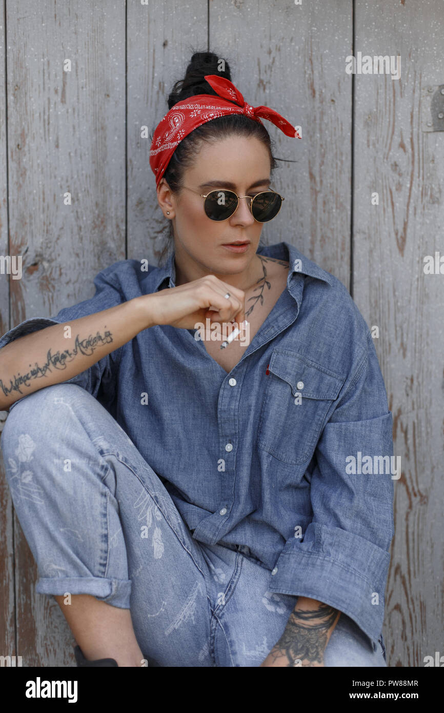 Cool American elegante hipster Mujer con gafas de sol en una camisa de  denim y jeans de moda con un cigarrillo sentada cerca de una vieja pared de  madera. Estilo pinup Fotografía