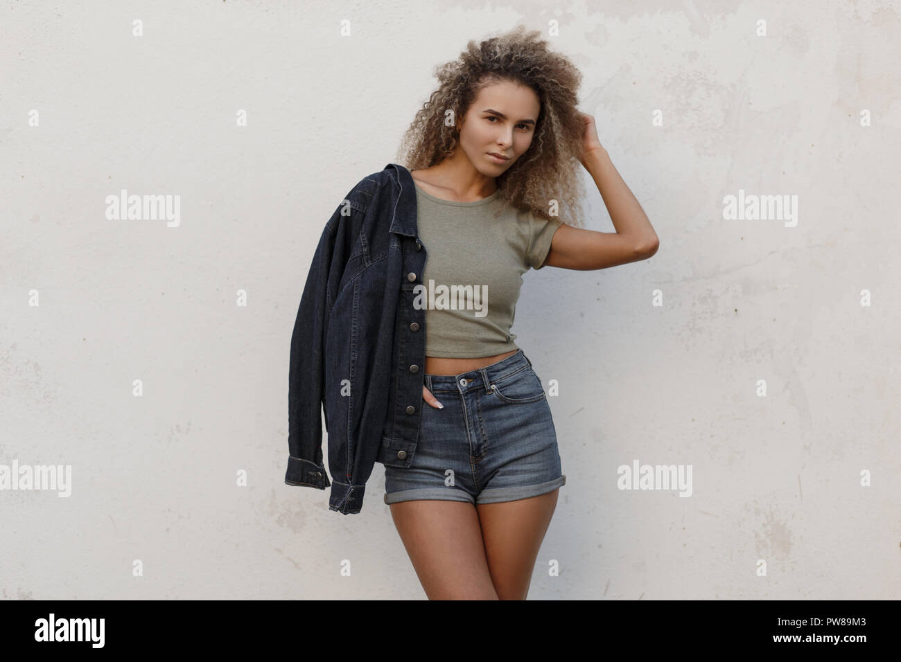 Elegante modelo joven y bella mujer con cabello rizado en moda denim jeans  ropa con una chaqueta y pantalones cortos de pie cerca de una pared blanca  en la playa Fotografía de