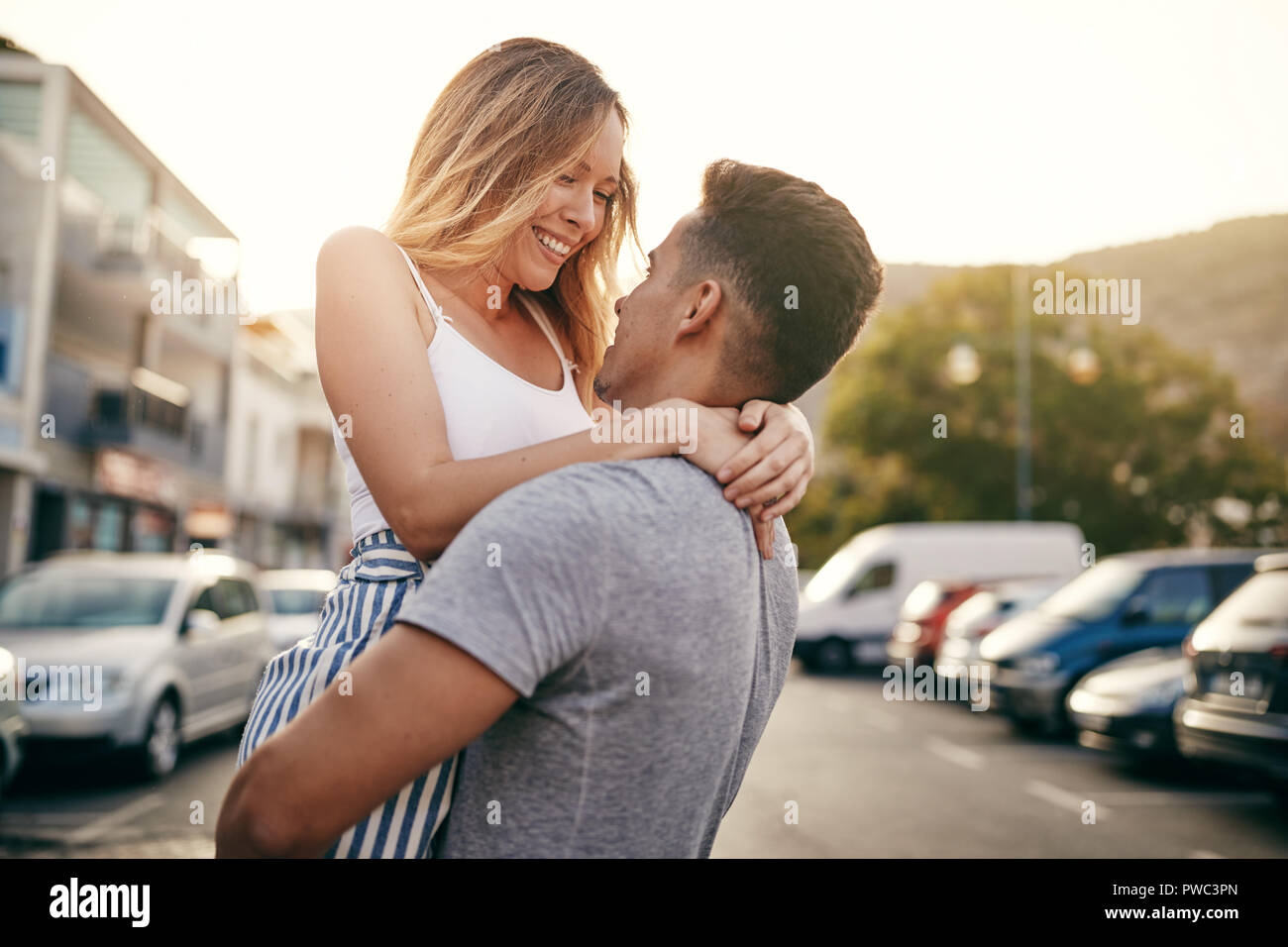 Joven sonriente sosteniendo a su novia en sus brazos mientras mirándose a los ojos en una calle de la ciudad al atardecer Foto de stock