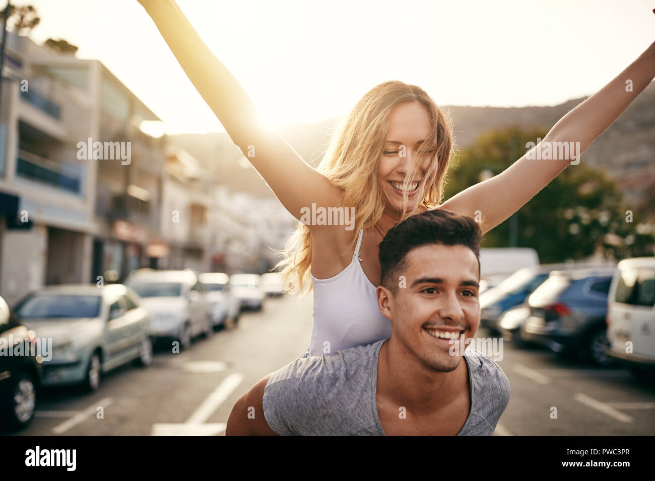 Laughing joven con sus brazos levantados siendo transportada sobre los hombros de su novio mientras se divierten juntos en la ciudad Foto de stock