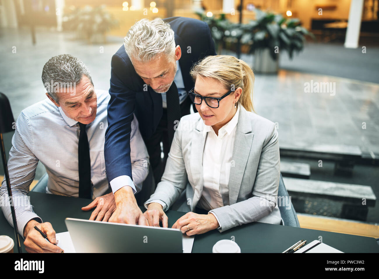 Administrador de maduro y dos compañeros de trabajo hablando juntos a través de un portátil durante una reunión en el vestíbulo de una moderna oficina Foto de stock