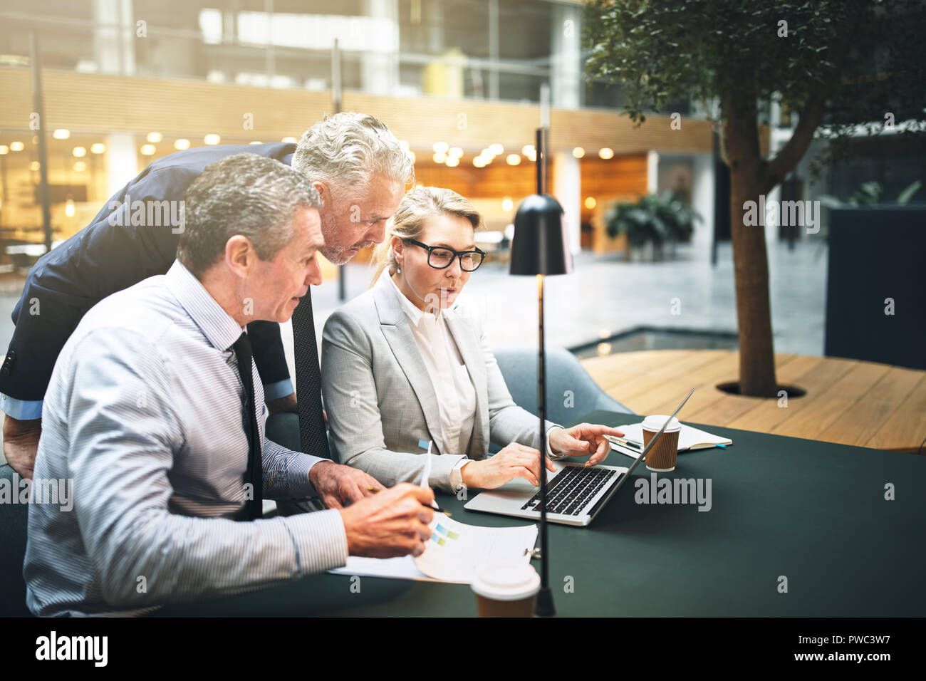 Tres ejecutivos maduros hablando juntos a través de un portátil mientras tiene una reunión en una mesa en el vestíbulo de un edificio de oficinas Foto de stock