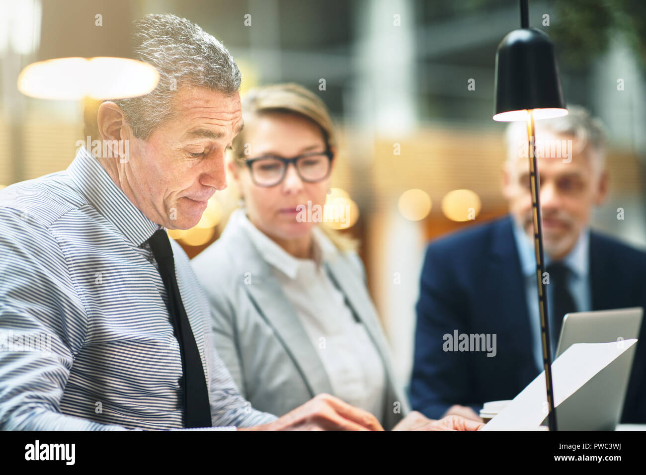 Empresario maduro leyendo papeleo durante una reunión con dos compañeros de trabajo mientras está sentado junto a una mesa en una oficina moderna Foto de stock