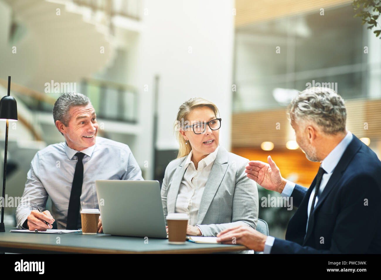 Tres compañeros de negocio maduro hablando de trabajar juntos, mientras que tener una reunión en una mesa en el vestíbulo de un moderno edificio de oficinas Foto de stock