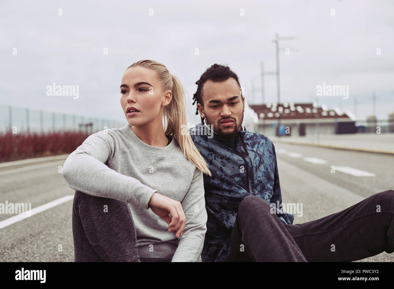 Diversos pareja joven sentada sobre una carretera en recuperar el aliento, mientras que para correr juntos en un día nublado Foto de stock