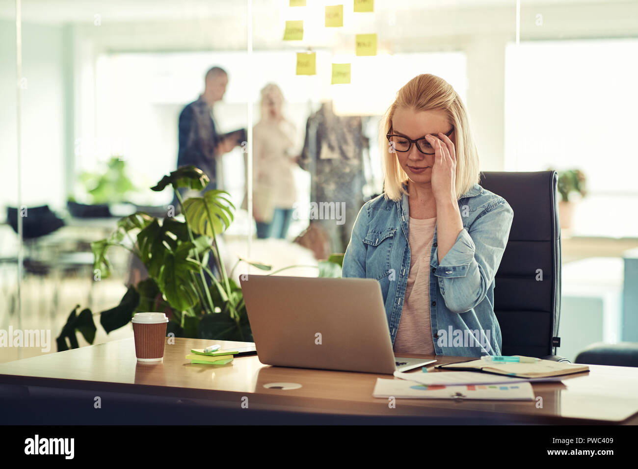 Vestida casualmente joven empresaria lleva gafas, concentrado en su trabajo mientras está sentado en un escritorio en una oficina moderna utilizando en un portátil. Foto de stock