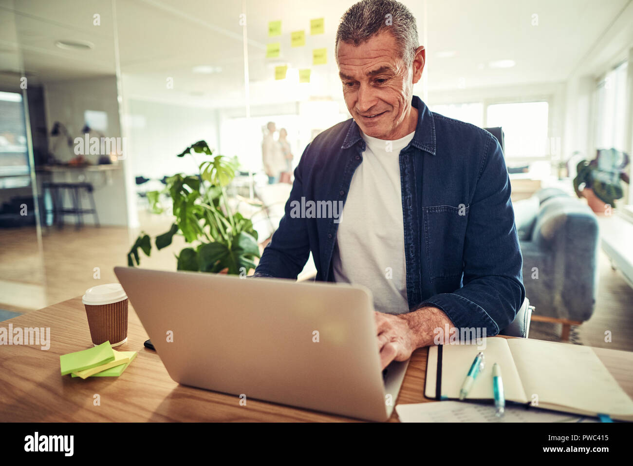 Vestida casualmente empresario maduro sonriendo mientras está sentado en un escritorio en su oficina de trabajo en línea con un portátil Foto de stock