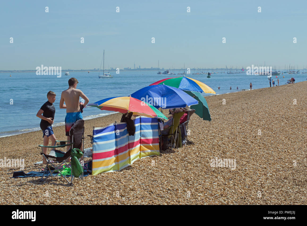 Cowes. La Isla de Wight. Pebble Beach. Costa. Los adultos bajo coloridas sombrillas y paraguas cortaviento. Dos chicos en natación atuendo. Barcos y personas. Recreación. Vacaciones. ​The Solent. Hampshire. ​England meridional. En el Reino Unido. Foto de stock