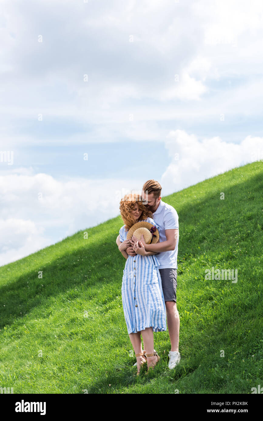 Pelirroja sonriente hombre abrace novia desde detrás de la colina verde Foto de stock
