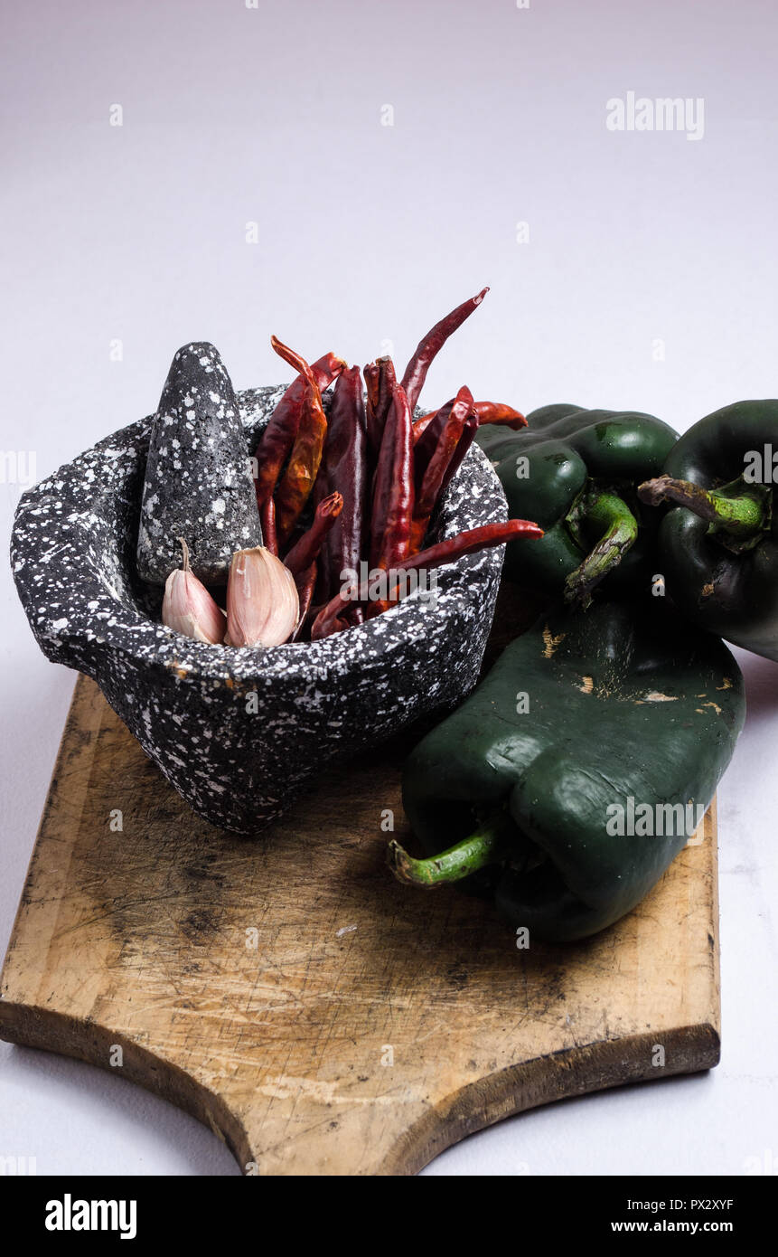 Mortero de piedra negra con los ingredientes para la salsa, pimientos rojos, ajo crudo, sobre una tabla de madera con quemaduras y grietas. Foto de stock