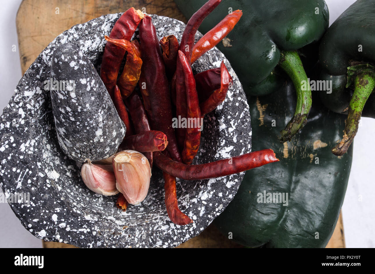 Mortero de piedra negra con los ingredientes para la salsa, pimientos rojos, ajo crudo, sobre una tabla de madera con quemaduras y grietas. Foto de stock