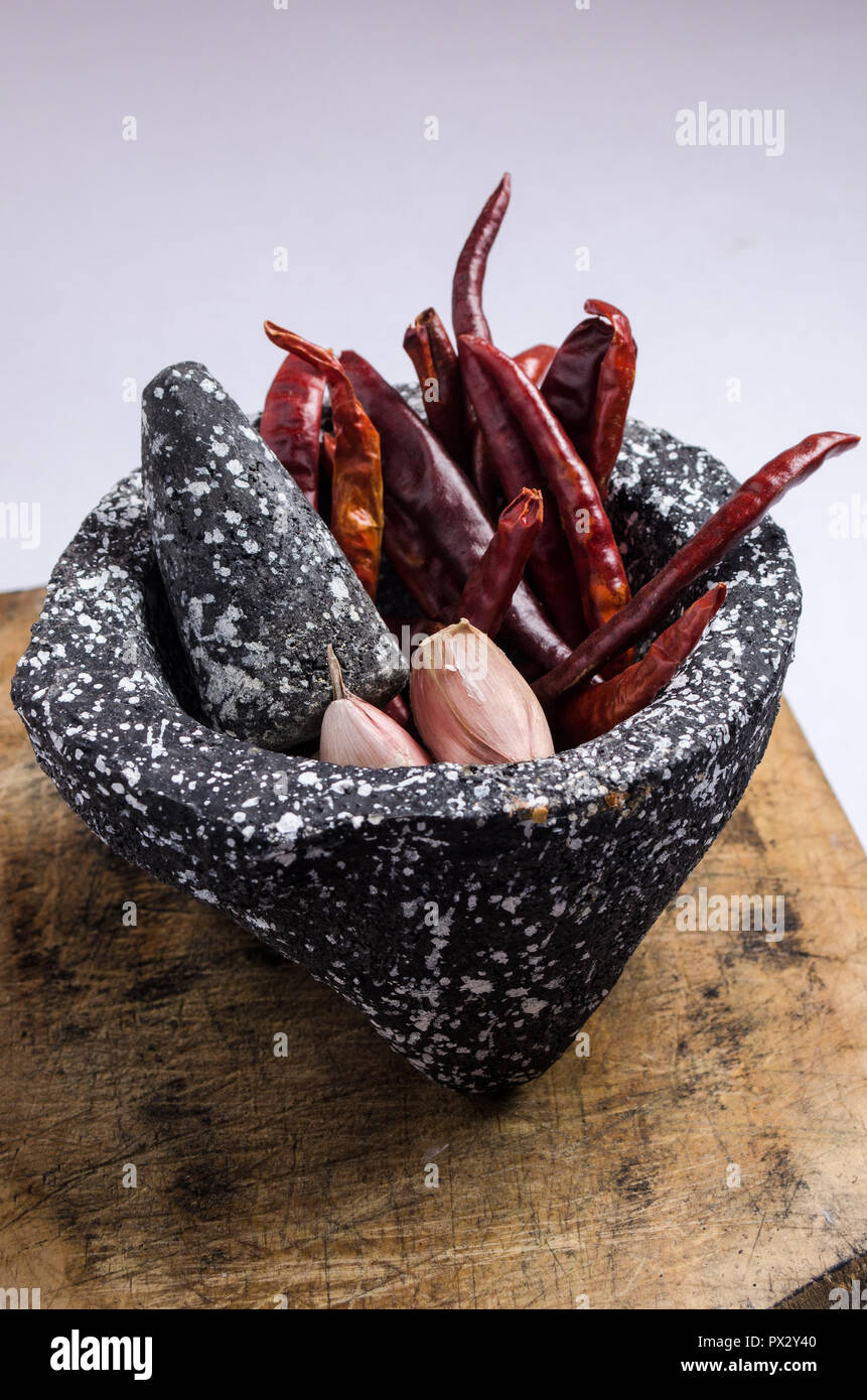 Mortero de piedra negra con los ingredientes para la salsa, pimientos rojos, ajo crudo, sobre una tabla de madera con quemaduras y grietas. Foto de stock