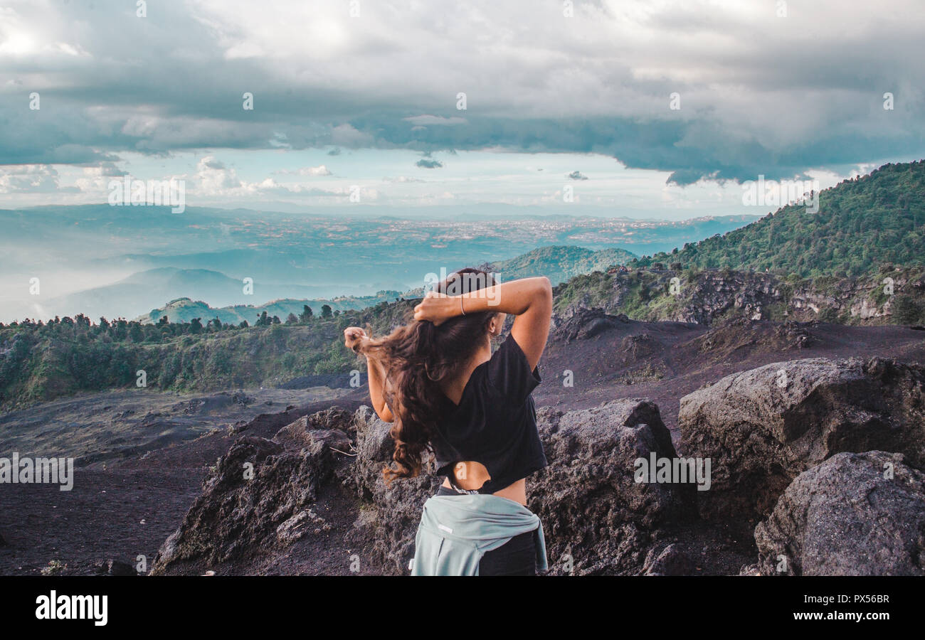 Mujer joven viajero gestos pelo en la parte superior del volcán Pacaya, mirando por encima de una roca volcánica y un exuberante valle en Guatemala Foto de stock