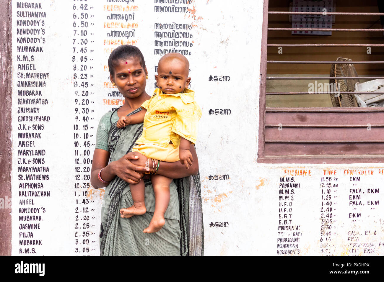 Mujer sosteniendo un bebé en la estación de autobuses de Kumily, Thekkady, Kerala, India. Ella está de pie delante de una lista de destinos. Foto de stock