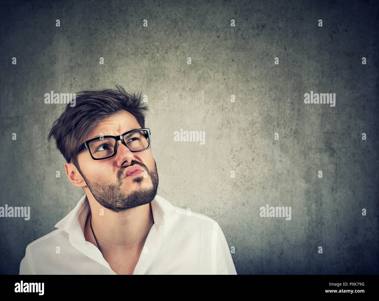 Adulto hombre barbado en vidrios y camisa blanca mirando en pensamientos siendo inciertas sobre fondo gris Foto de stock