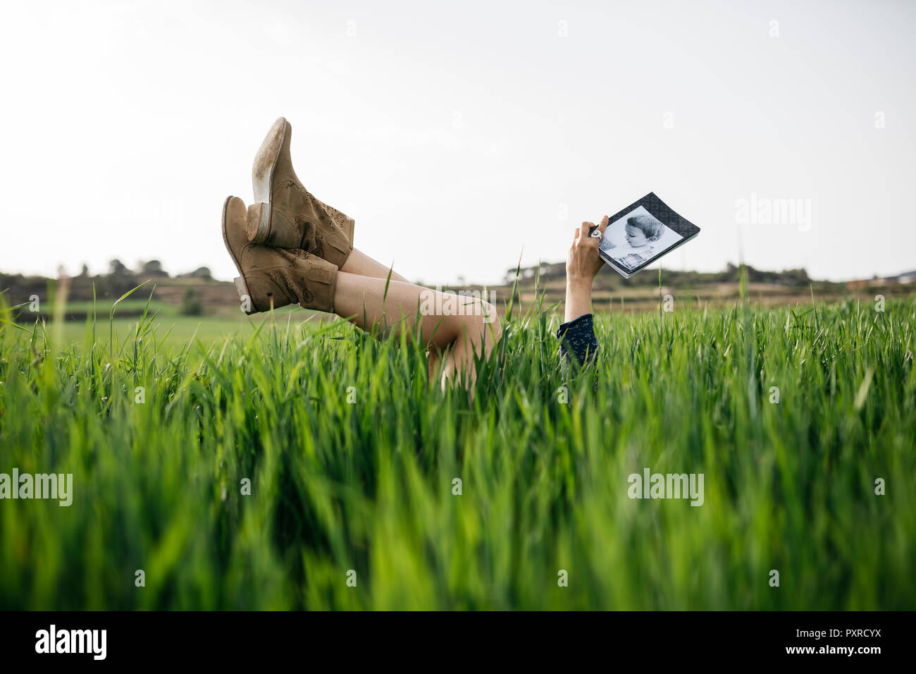 Mujer recostada en un campo con las piernas en el aire leyendo un libro Foto de stock
