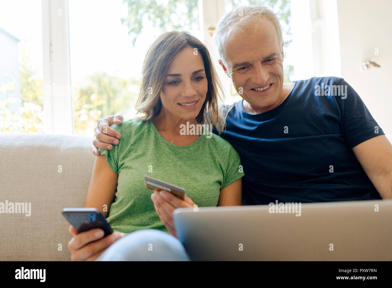 Pareja sonriente sentado en el sofá en casa de compras online con smartphone laptopand Foto de stock