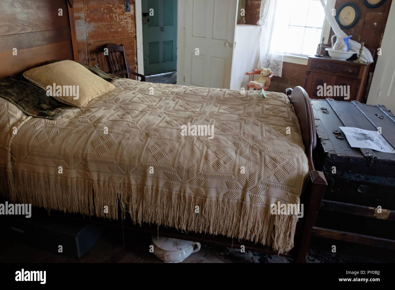 Cama antigua cubierta en la cama con almohadas y viejo tronco de madera rústica al pie de la cama. Interior de una casa en la aldea histórica plaza de castaños, McKinney, Texas. Foto de stock