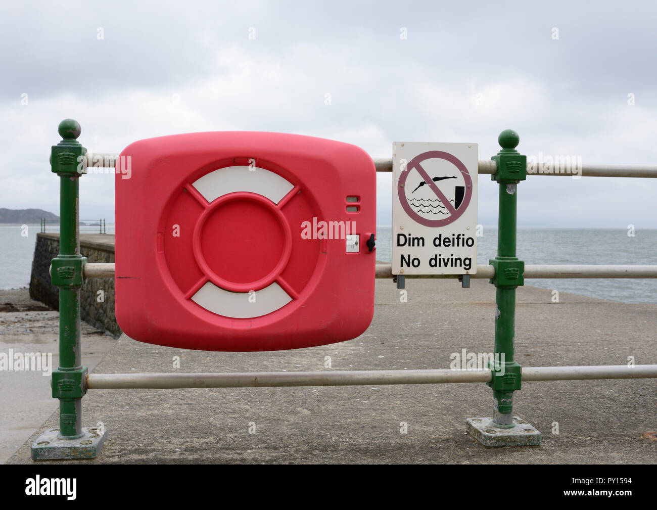 Caja de salvavidas y bilingüe en inglés y galés no buceo signo fijo a  barandilla en muelle de concreto, con vistas al mar en Criccieth al norte  de Gales, Reino Unido Fotografía