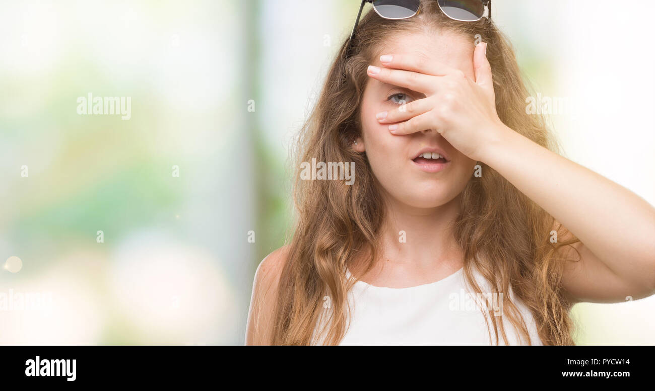 Joven Mujer rubia con gafas de sol peeking en shock que cubre el rostro y  los ojos con la mano, mirando a través de los dedos con expresión  avergonzada Fotografía de stock -