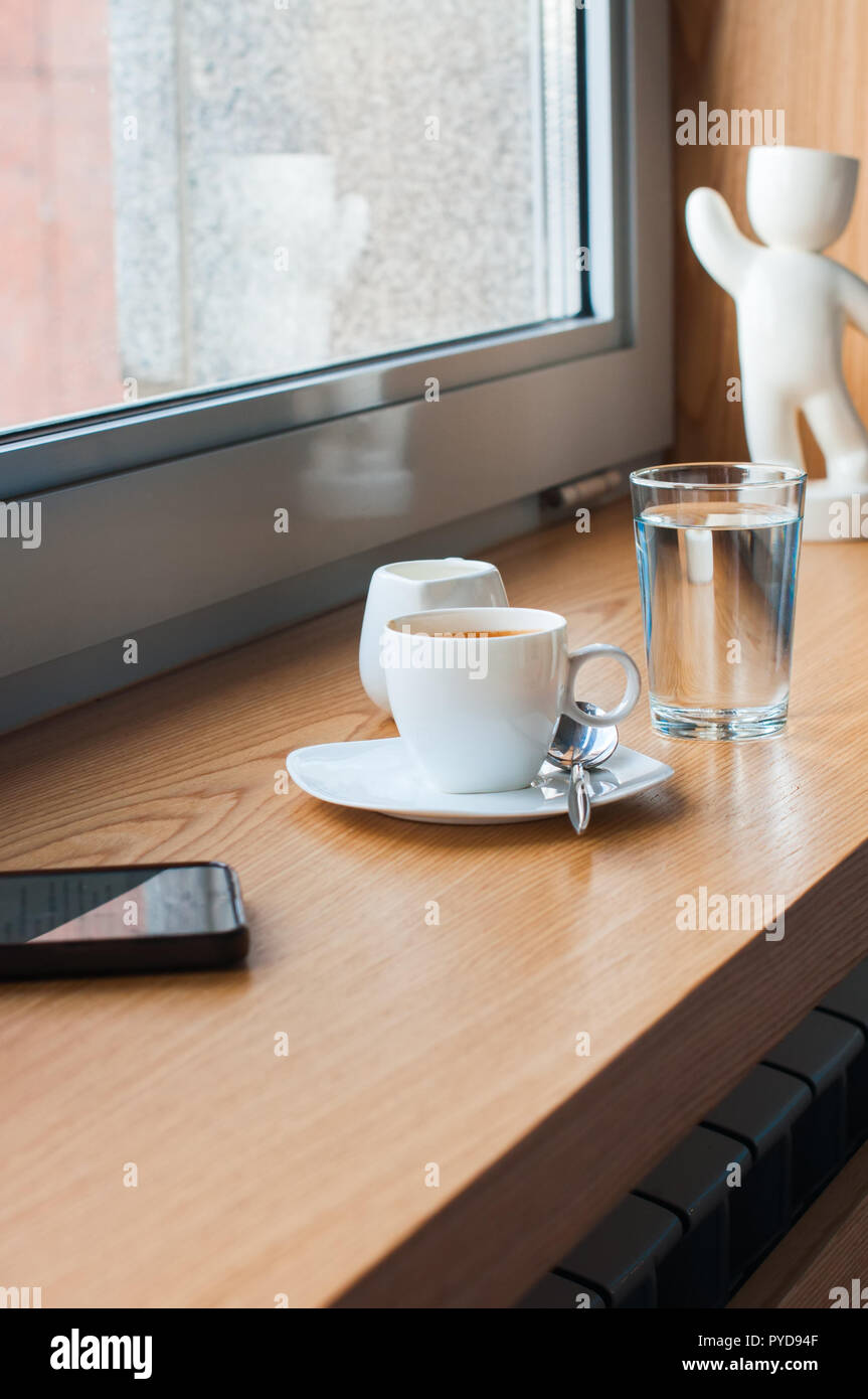 Taza de expreso, vaso de agua, teléfonos inteligentes, leche en una ventana Foto de stock