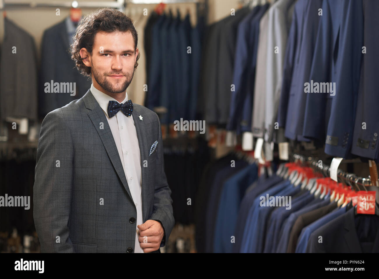 Retrato de hombre elegante traje gris con pajarita y camisa blanca posando  en cámara. Guapa morenita con barba sonriente, sosteniendo la mano en el  bolsillo. Los estantes con camisas en segundo plano