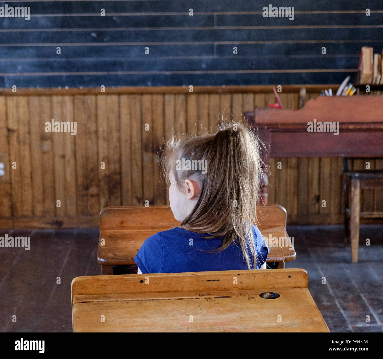 Niña de la escuela en la parte superior azul se sienta en vintage escritorio de madera en el aula. Wilmeth Schoolhouse, plaza de castaños Pueblo Histórico, McKinney Texas. horizontal. Foto de stock