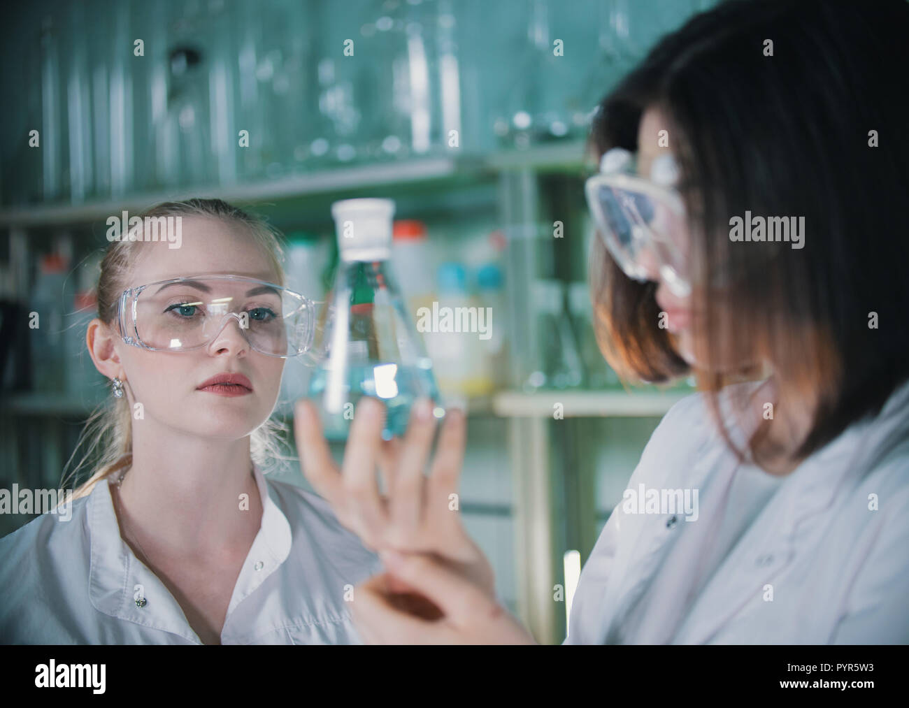 Dos jóvenes mujer sosteniendo un laboratorio químico en frascos con líquido claro en él. Mujer rubia en foco Foto de stock