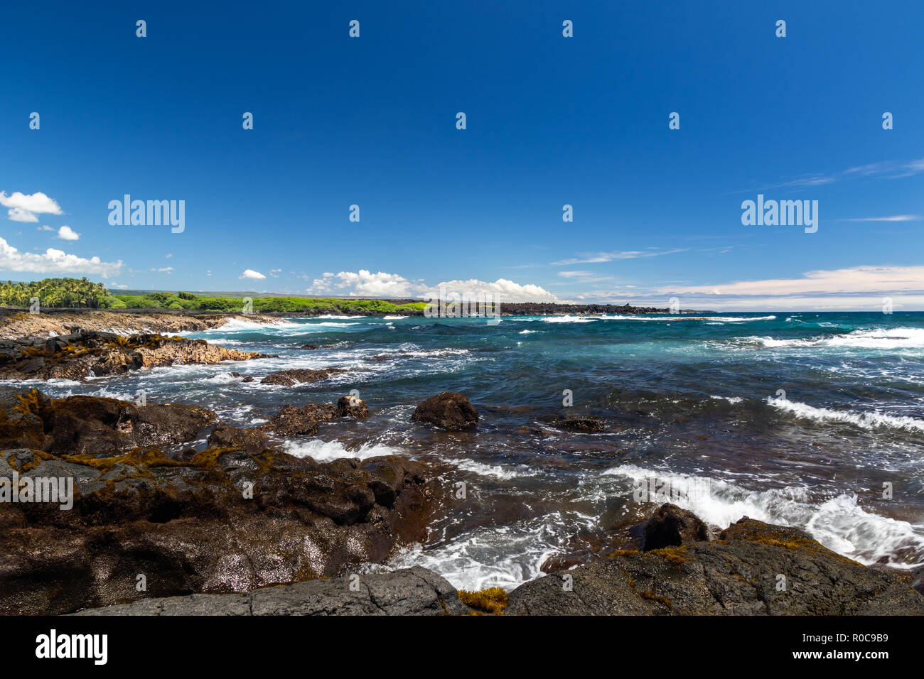 Vista al mar en la playa de arena negra en la isla más grande de Hawai. Océano Pacífico azul con olas, áspera roca volcánica y vegetación marrón en la orilla. Foto de stock
