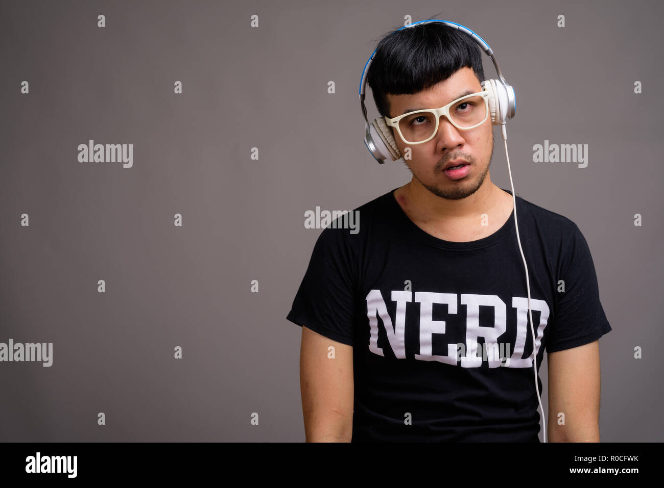 Joven asiático nerd hombre escuchando música contra el fondo gris Foto de stock