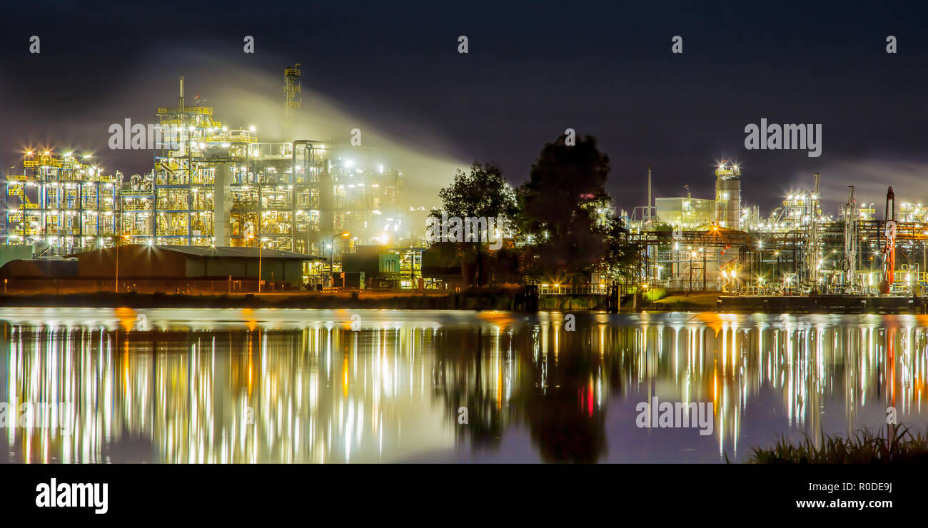 Panorama de la escena nocturna de detalle de una planta industrial química pesada reflejando en el agua con mazework de tubos en crepúsculo Foto de stock