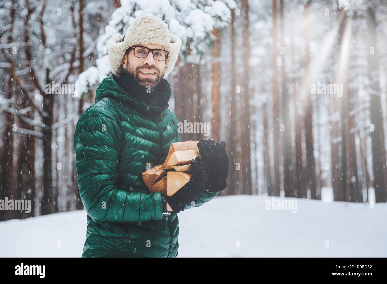 Unshaven sonriendo macho posee pila de leña, va a hacer fuego para calentar a sí mismo en el periodo de heladas, pasan el invierno en el bosque, el día tiene un picnic con un amigo Foto de stock