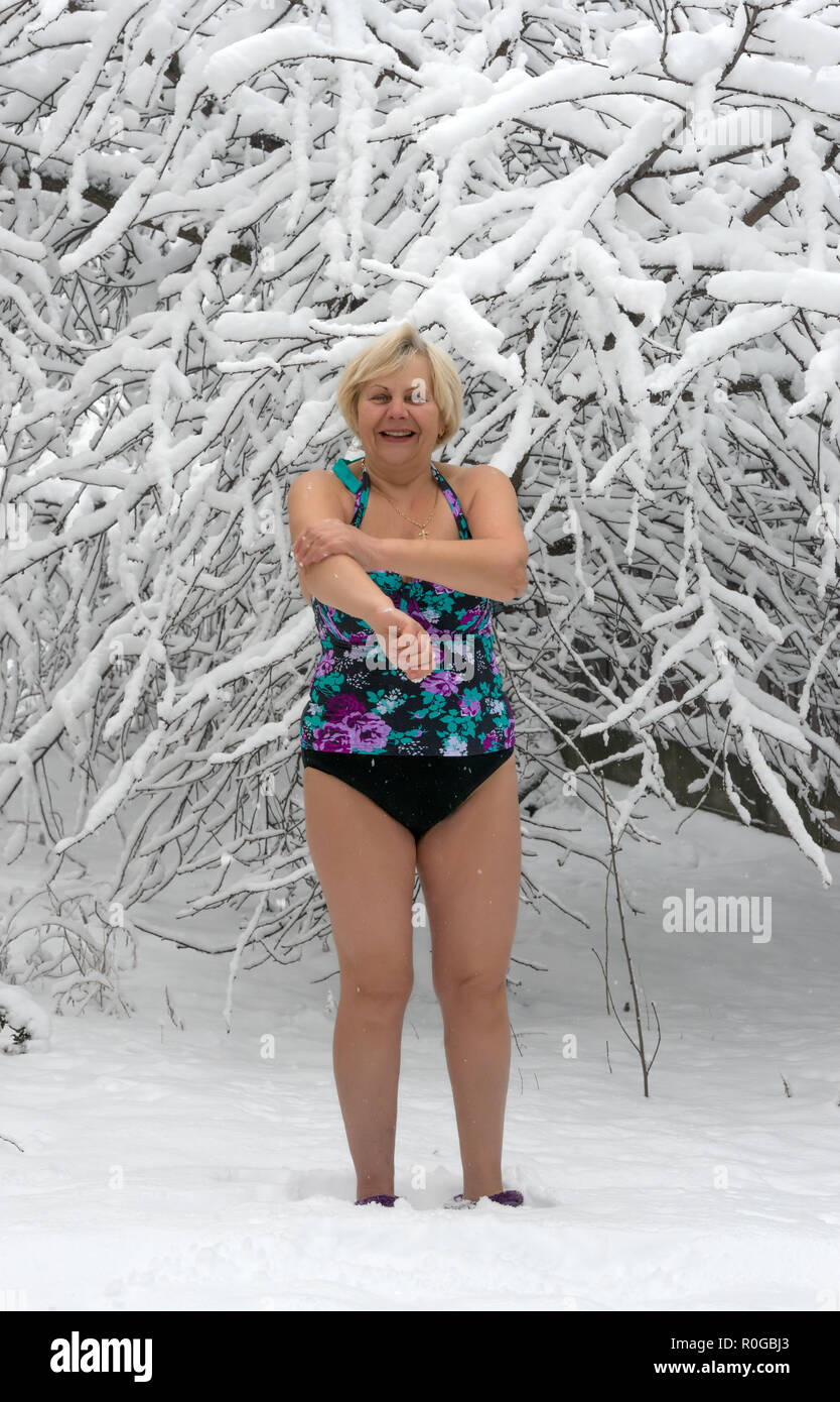 Retrato de mujer senior curtidos en traje de baño está de pie sobre la nieve  fresca en el jardín cubierto de nieve cerca de cerezo y agarrándose a sí  misma con nieve