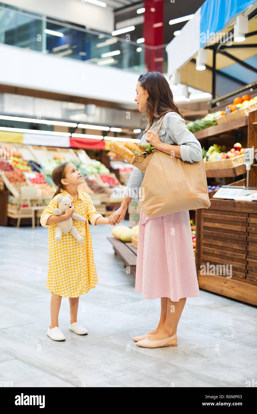 Sonriente joven madre en ropa informal mantener la bolsa de comida orgánica  y caminar con mi hija en el mercado de los granjeros, ellos conversando y  tomándose de la mano Fotografía de