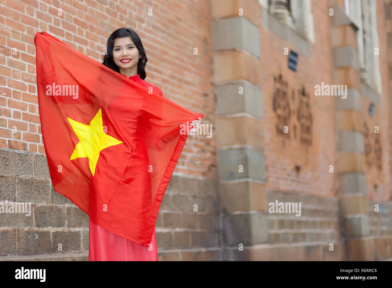 Patriótica feliz joven vietnamita con una encantadora sonrisa sosteniendo la bandera nacional en sus brazos como ella se levanta contra edificios de ladrillo en una u Foto de stock