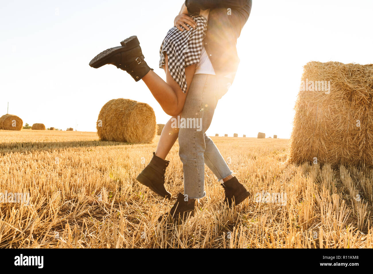 Foto recortada de chico y chica enamorada caminando a través de campo de oro con montón de montones de heno y abrazando durante el día soleado Foto de stock