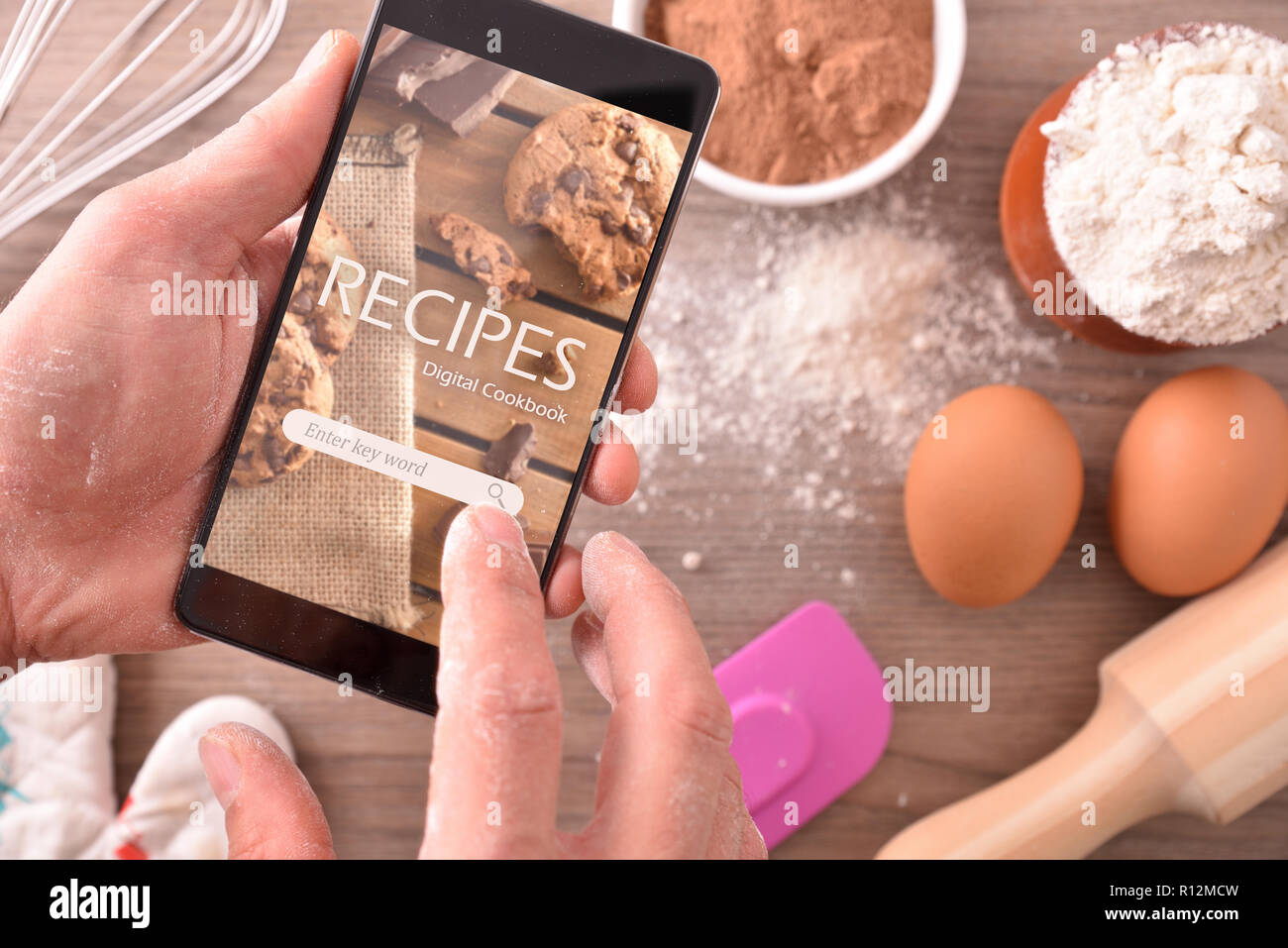 Manos con mobile app en recetas con ingredientes de pastelería de fondo. Usando el smartphone a cocinar. Concepto de recetas en libro digital. Composit horizontal Foto de stock