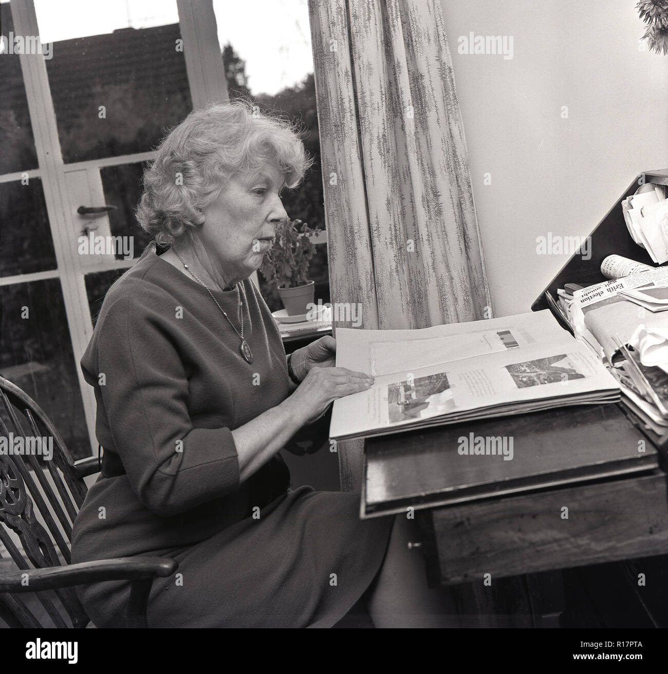 1965, una madura señora sentada en el interior de una habitación en un escrito de una mesa o escritorio mirando a través de un álbum de fotos personales, revivir los recuerdos de su vida. Foto de stock