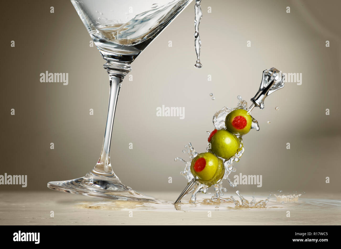 Imagen recortada de copa de martini salpicaduras de líquido y las aceitunas sobre la superficie, fondo gris Foto de stock