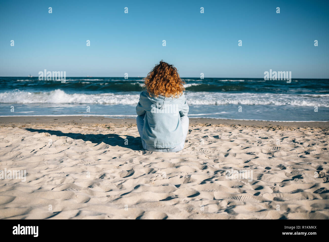 Mujer joven sentado en la playa mirando las olas, vista trasera. Mujeres con cabello rizado rojo vestidos en denim jacket descansando en la orilla del mar en sunny d Foto de stock