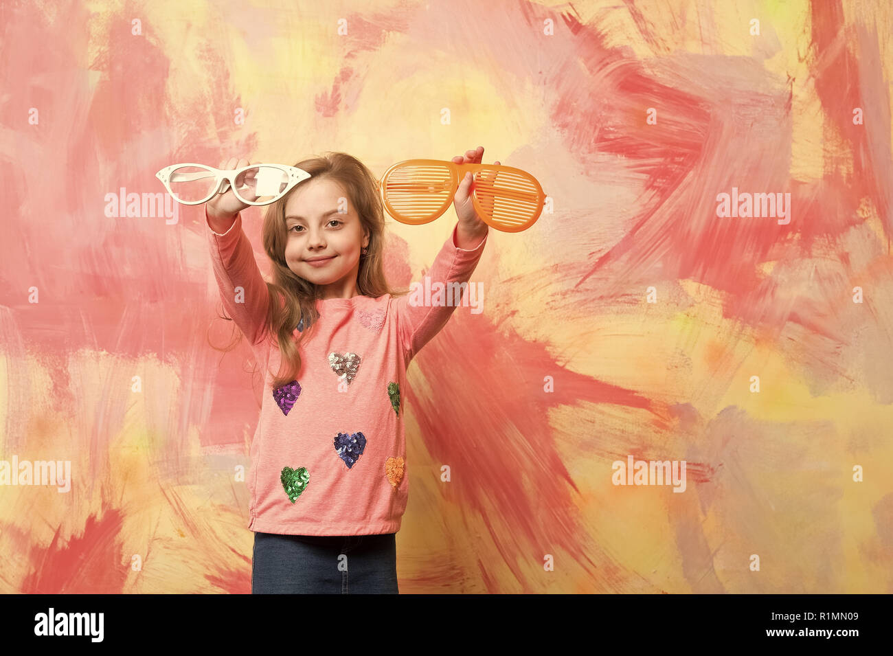 Chica sonriente con dos funny gafas de sol. Modelo de bebé, con un largo  cabello rubio en ropa de moda en colorido mural abstracto. Concepto de moda  infantil. Infancia feliz Fotografía de