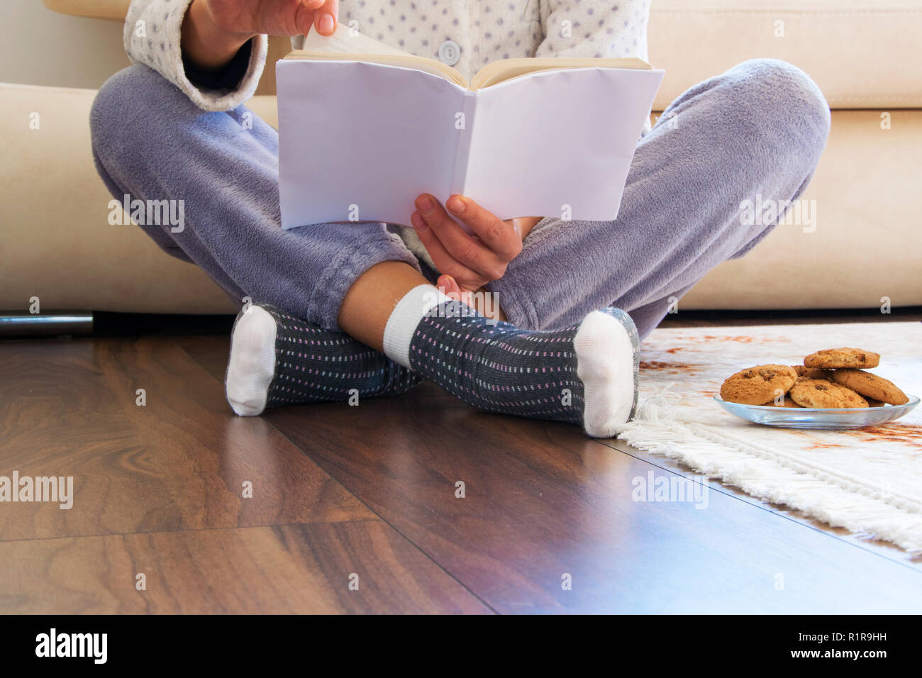 Vista recortada de mujer con pijamas sentado en el piso de madera en casa  con bebidas calientes y galletas, leyendo un libro- acogedor del concepto  de la vida cotidiana Fotografía de stock -