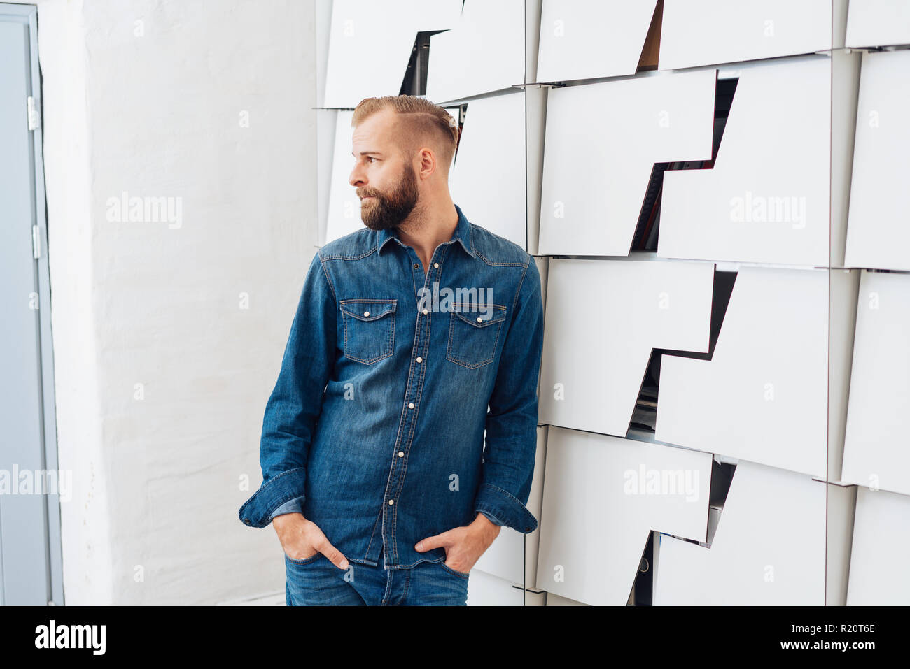 Joven Hombre Barbado Con Un Elegante Corte De Pelo Corto Vestido Con Jeans Y Jeans Camisa De Pie Junto A La Pared Gris Decorativo Con Las Manos En Los Bolsillos Y Fotografia