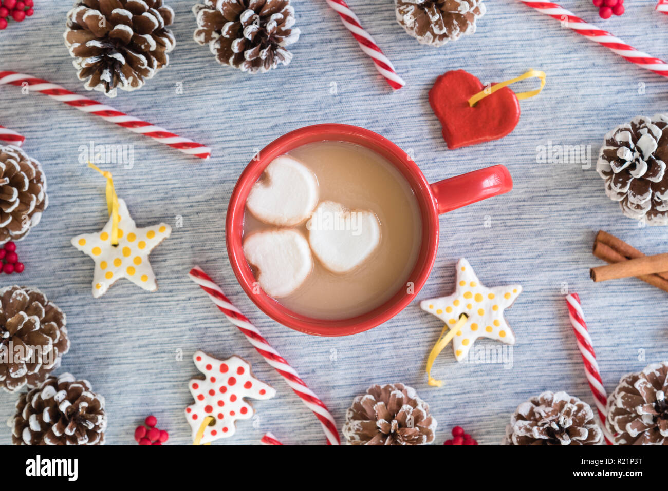 Chocolate caliente en taza roja con corazón malvaviscos rodeado de adornos artesanales, bastones de caramelo y piñas Foto de stock