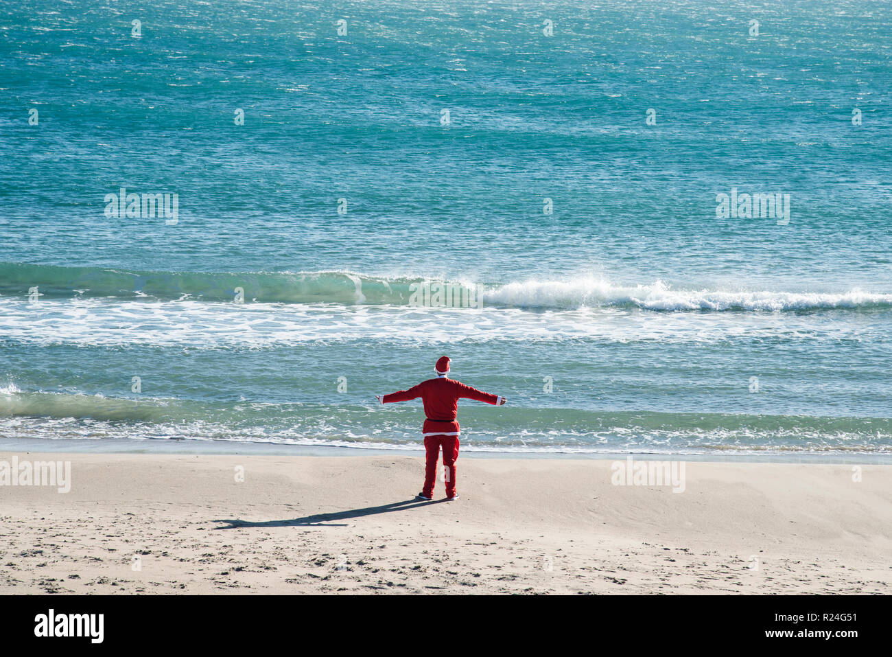 Joven vistiendo un traje de Papá Noel, visto desde detrás, de pie en la playa frente al océano, con sus brazos extendidos en el aire Foto de stock