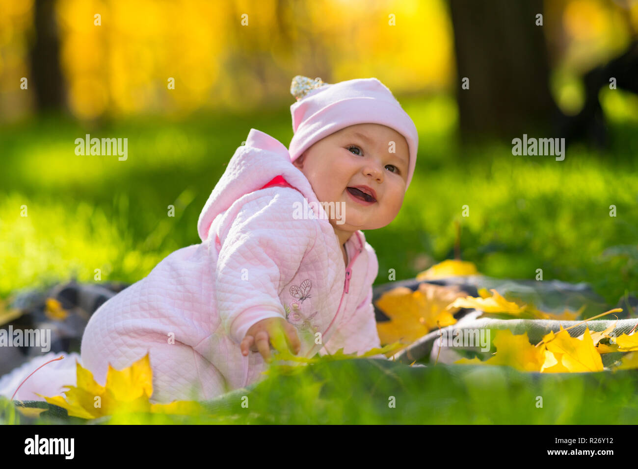 Feliz riendo hijita en un traje rosa acostada sobre una manta sobre el exuberante césped verde en un parque en otoño con coloridas hojas Foto de stock