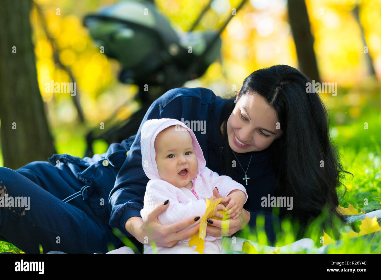 Feliz a la madre y al bebé niña jugando en un parque en una cobija en el pasto verde jugando con coloridas hojas en otoño de árboles circundantes Foto de stock
