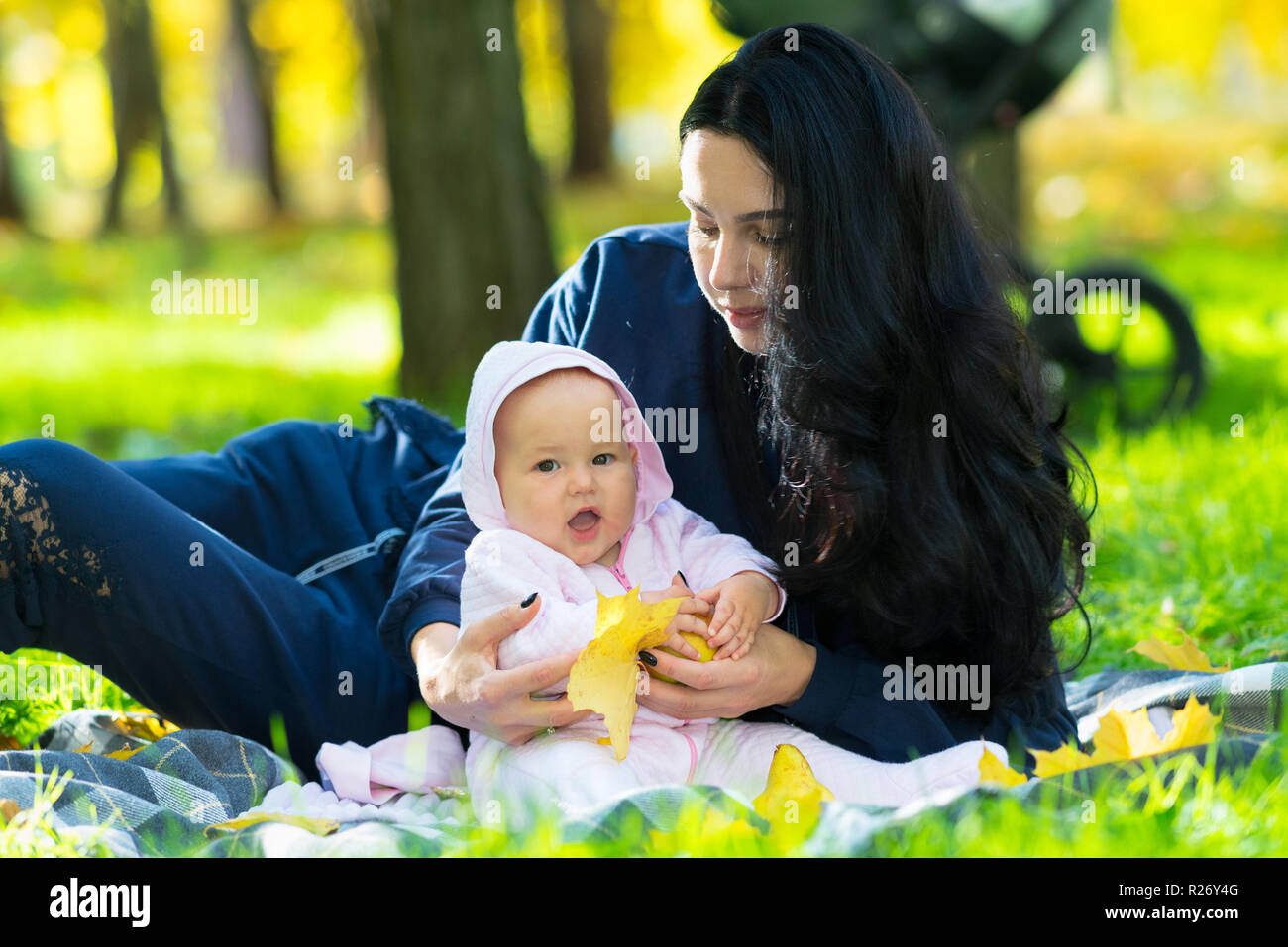 Riendo poco feliz bebé niña jugando con un colorido amarillo hojas caer como ella lo abraza con su amorosa madre sobre una manta en el parque Foto de stock
