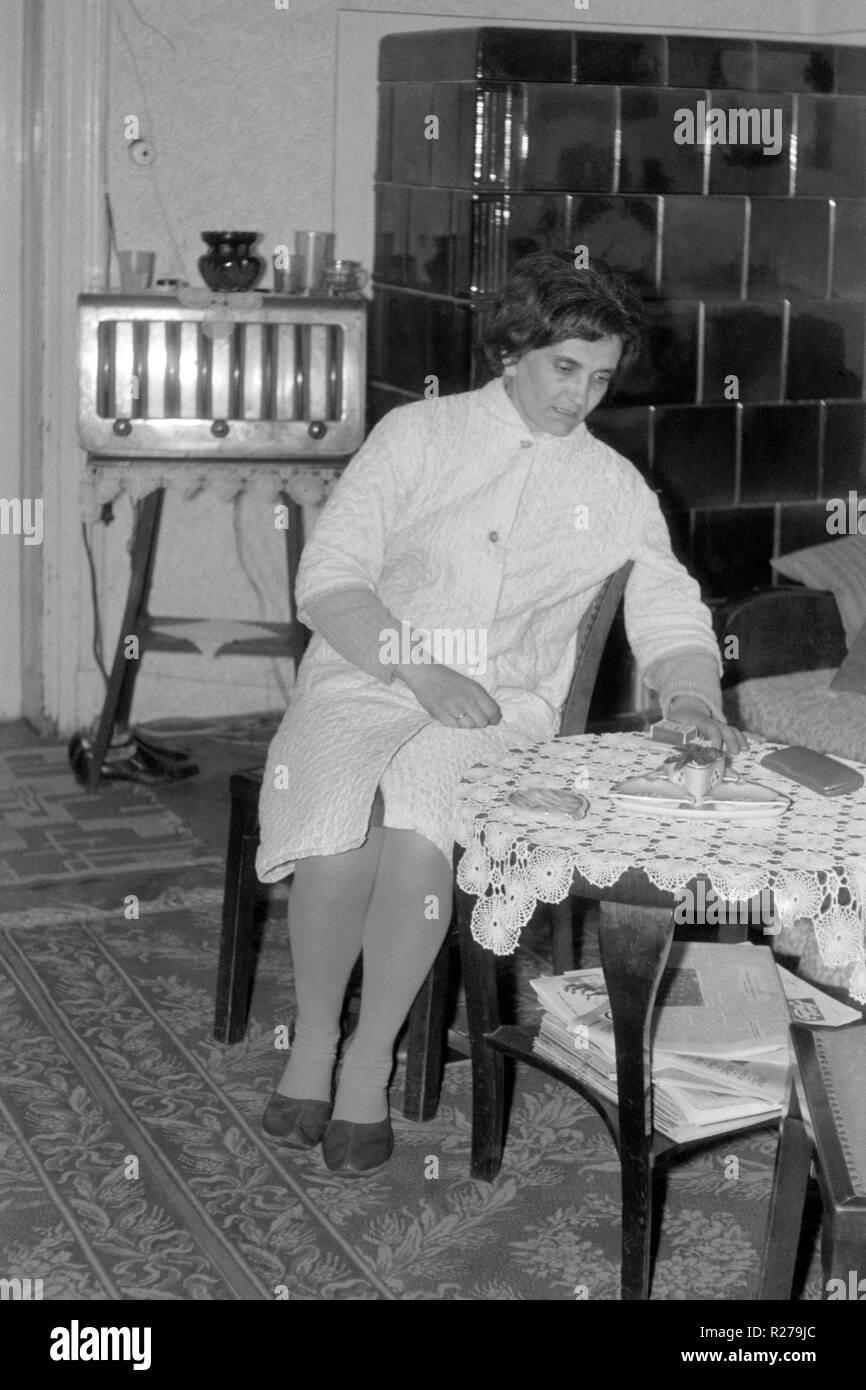 Hembra madura sentado en la sala de estar en la mesa con el vintage radio y gran chimenea de cerámica en el fondo de 1960 Hungría Foto de stock